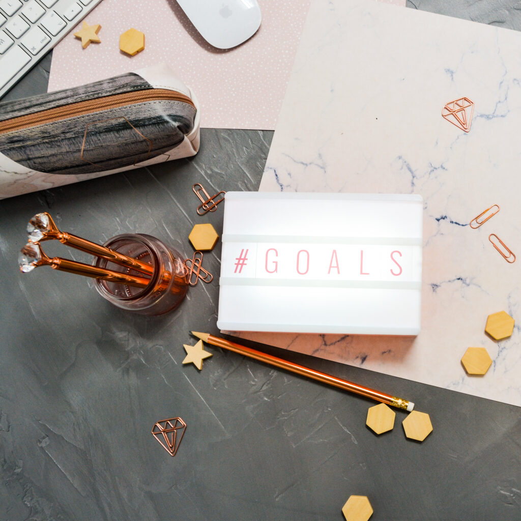 dark-grey-background-with-scattered-pink-papers-paperclips-and-wooden-charms-Sticky-notepad-pencil-case-silver-keyboard-and-mouse-scaled