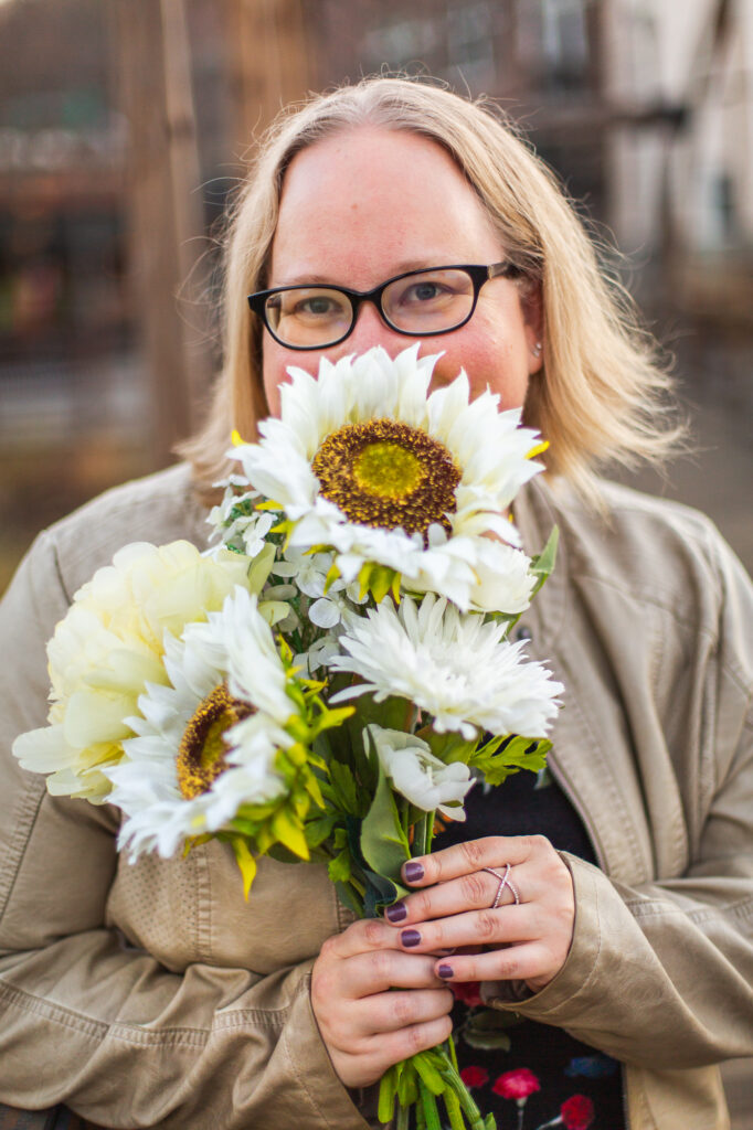 headshot of blond woman wearing black glasses smelling sunflowers