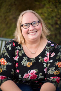 headshot of blond woman wearing black glasses sitting on a bench