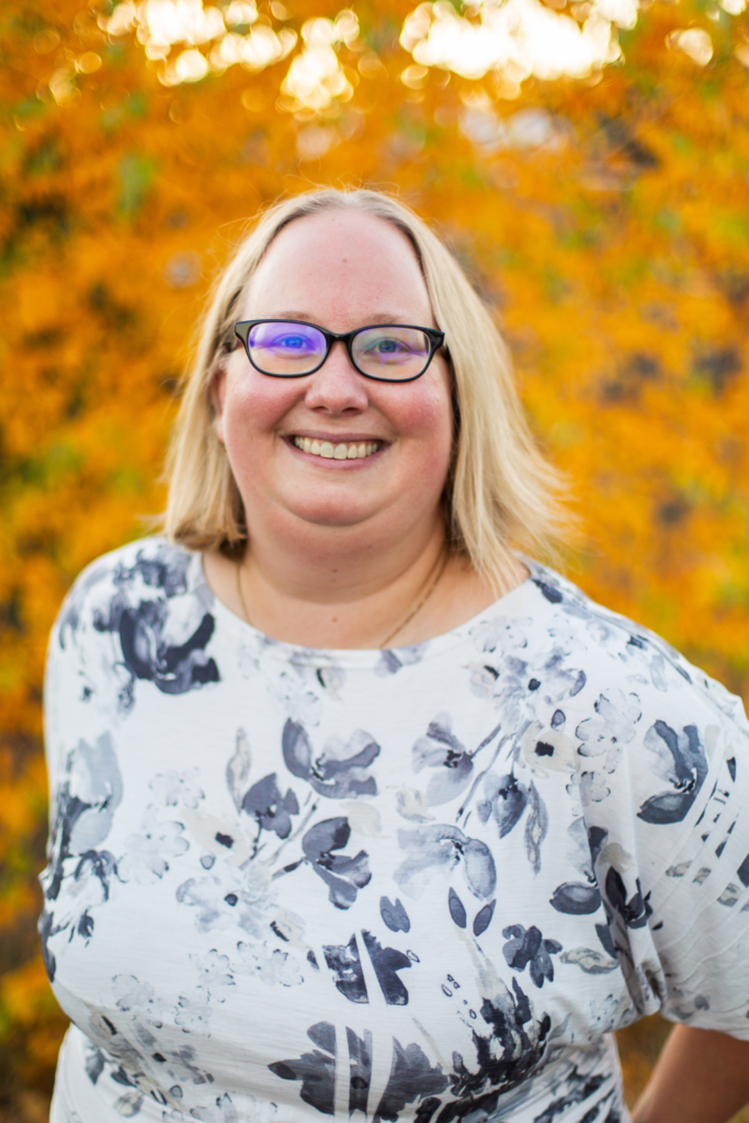headshot of blond woman wearing black glasses in front of a tree with yellow leaves
