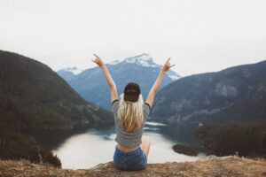 girl with arms raised sitting on hill overlooking alpine lake and mountains