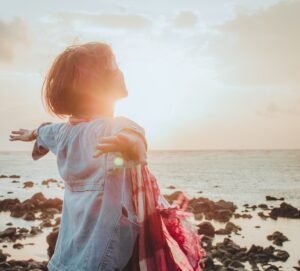 Girl in front of rocky ocean shore with arms extended in sunshine