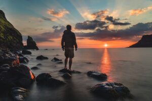 person standing on rocks looking at sunset reflection in ocean