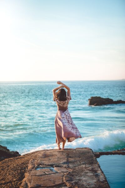 woman with arms raised looking out at blue ocean and waves
