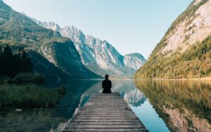 Girl sitting on a wooden dock looking at a calm lake and mountains on a clear day
