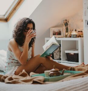 Girl sitting on bed drinking from a mug reading a book
